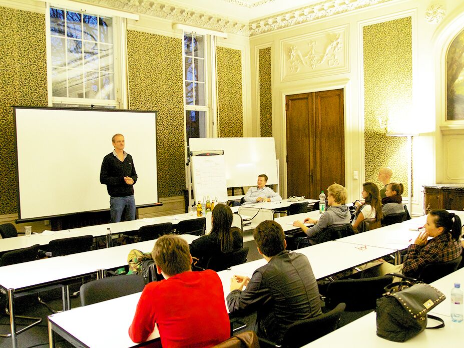 Ein Mann hält einen Vortrag vor einer Gruppe Studierender in einem prunkvoll gestalteten Seminarraum mit Leinwand und Flipchart.