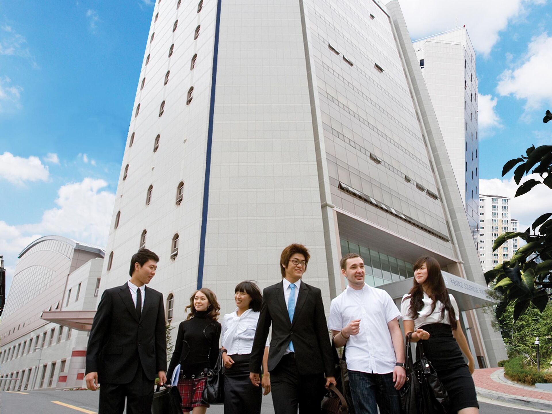 Sechs junge Menschen in Businesskleidung gehen lachend vor einem hohen modernen Gebäude.