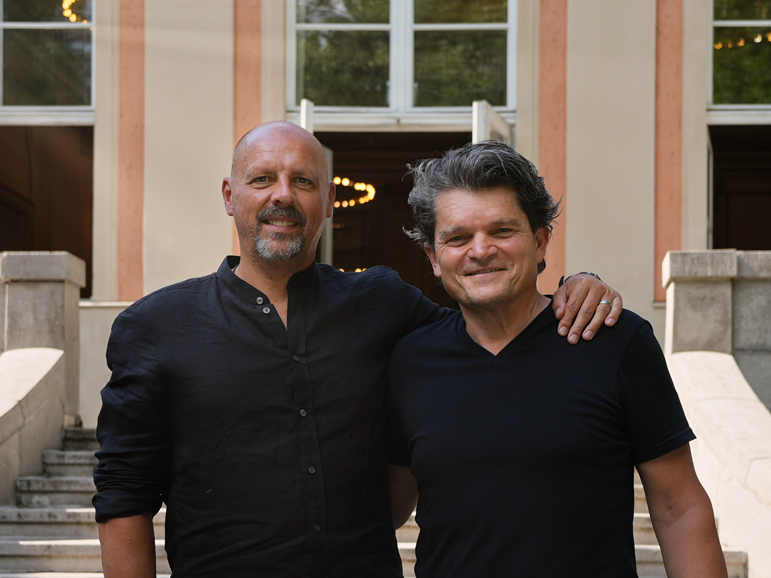 Zwei Männer stehen lächelnd nebeneinander vor einem Gebäude. Einer hat den Arm um die Schulter des anderen gelegt.