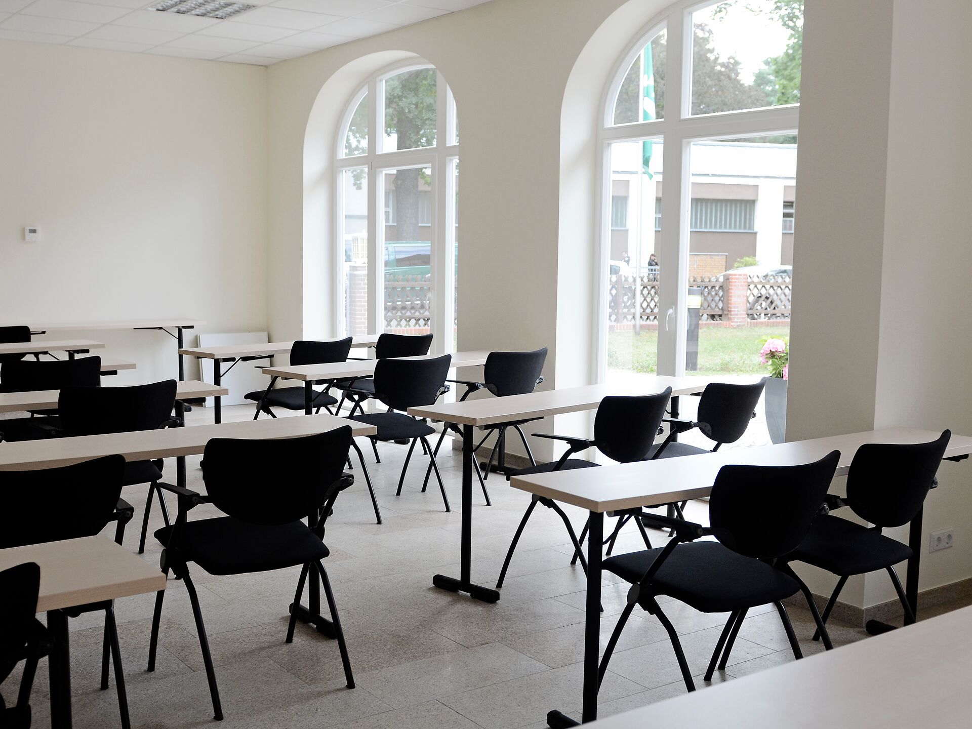 Heller Seminarraum mit großen Fenstern, leeren Tischen und schwarzen Stühlen.
