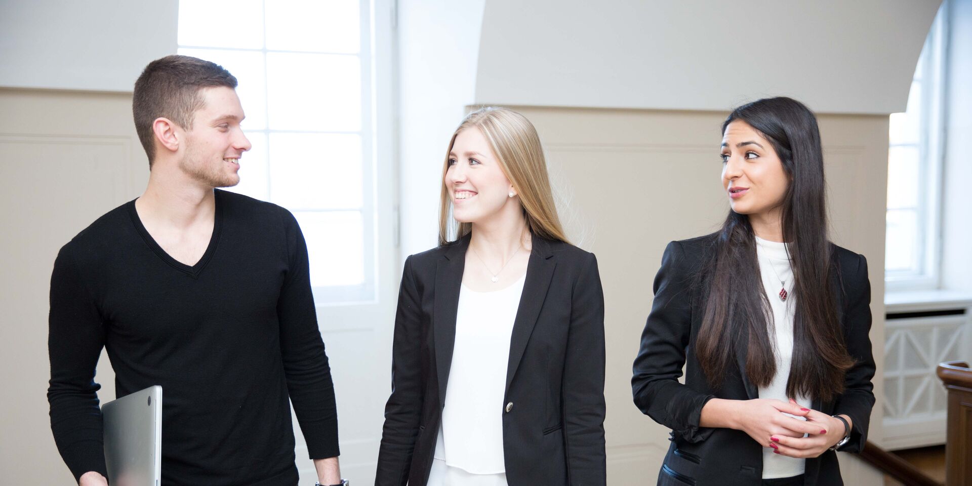 Drei junge Menschen im Business-Outfit gehen gemeinsam durch einen hellen Raum und unterhalten sich.