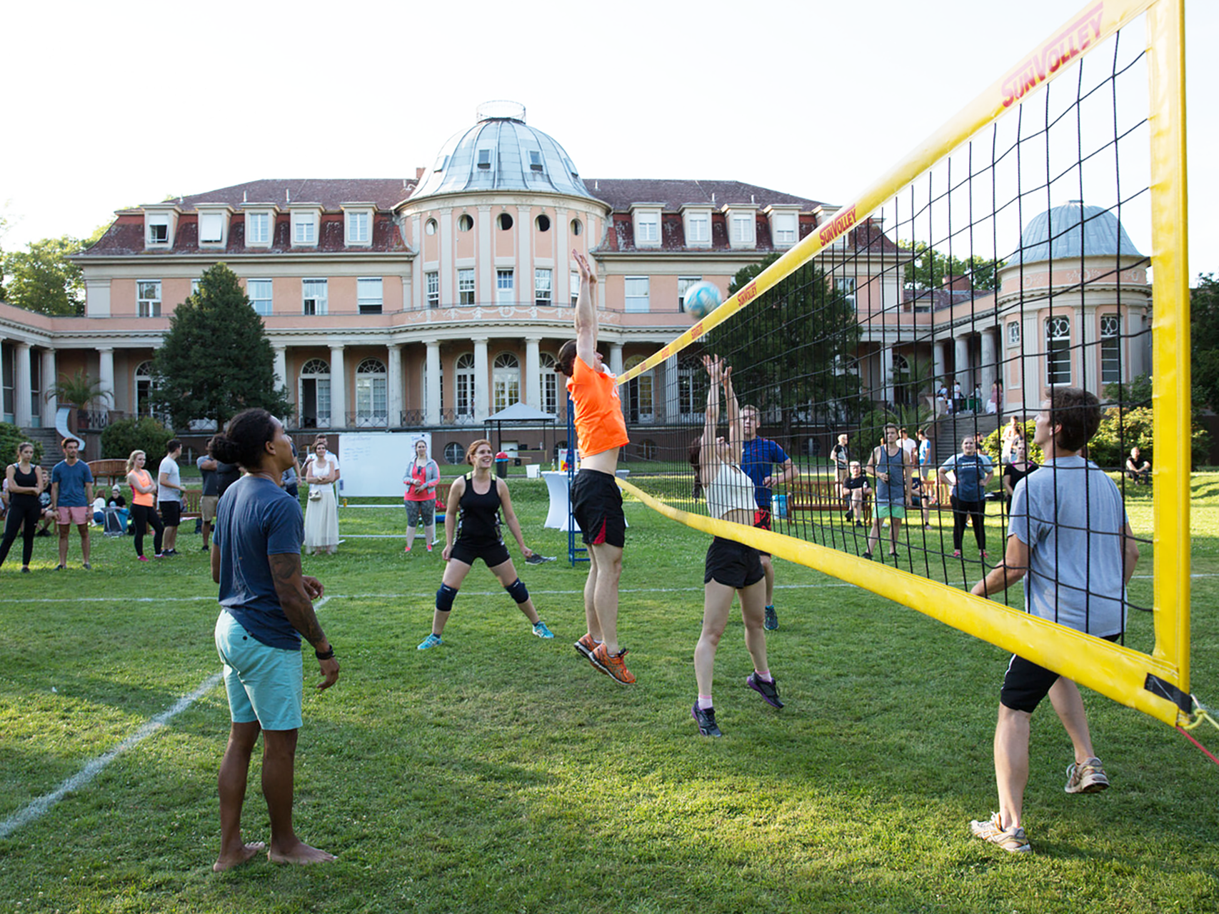 Eine Gruppe von Menschen spielt Volleyball im Freien auf einer Wiese vor einem großen historischen Gebäude. Ein Spieler in orangem Shirt springt hoch, um den Ball über das Netz zu schlagen, während Gegenspieler versuchen, ihn zu blocken. Zuschauer stehen