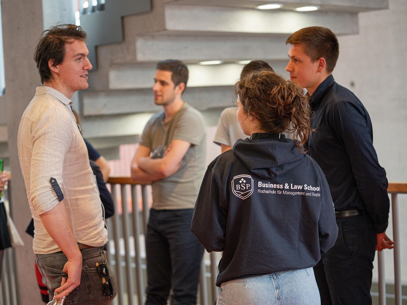 Mehrere junge Menschen stehen in einem Gebäude und unterhalten sich, eine Person trägt einen schwarzen Hoodie mit dem Logo der BSP Business & Law School.
