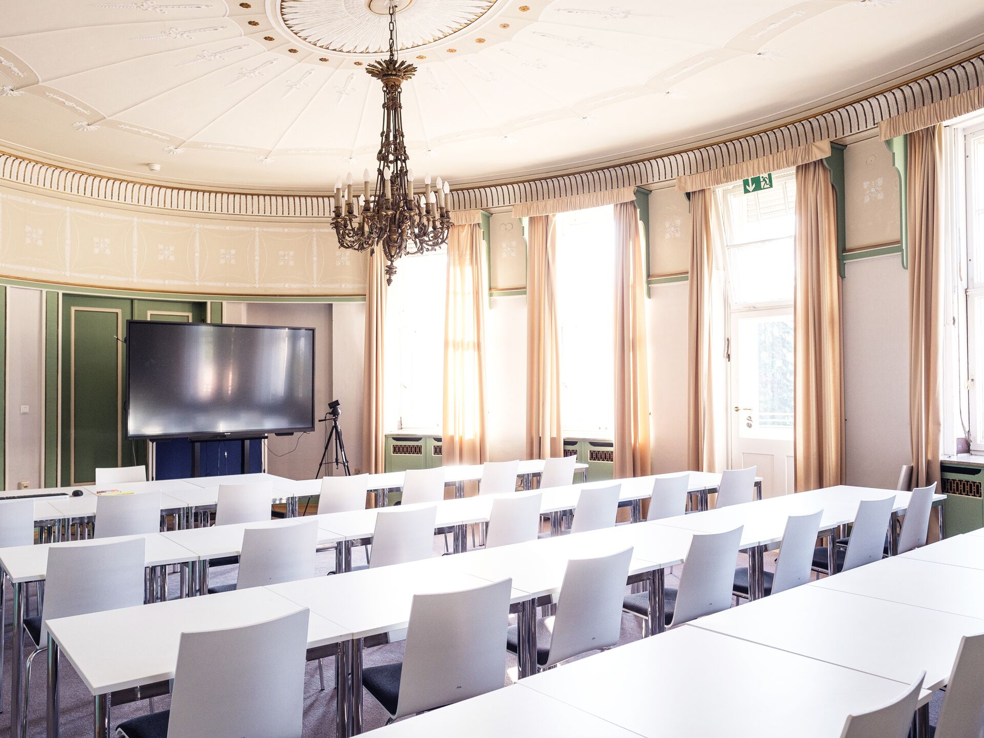 Ein heller Seminarraum mit weißen Tischen und Stühlen, großen Fenstern mit Vorhängen und einem Kronleuchter an der Decke. Vorne steht ein großer Bildschirm neben einem aufgestellten Stativ mit Kamera.