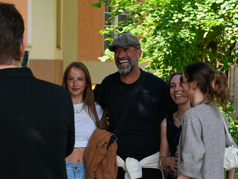 Eine Gruppe von Menschen steht draußen zusammen und lacht. Ein Mann mit Schirmmütze und Bart steht in der Mitte, neben ihm junge Frauen. Im Vordergrund ist eine Person mit Smartphone zu sehen, die offenbar ein Foto macht.