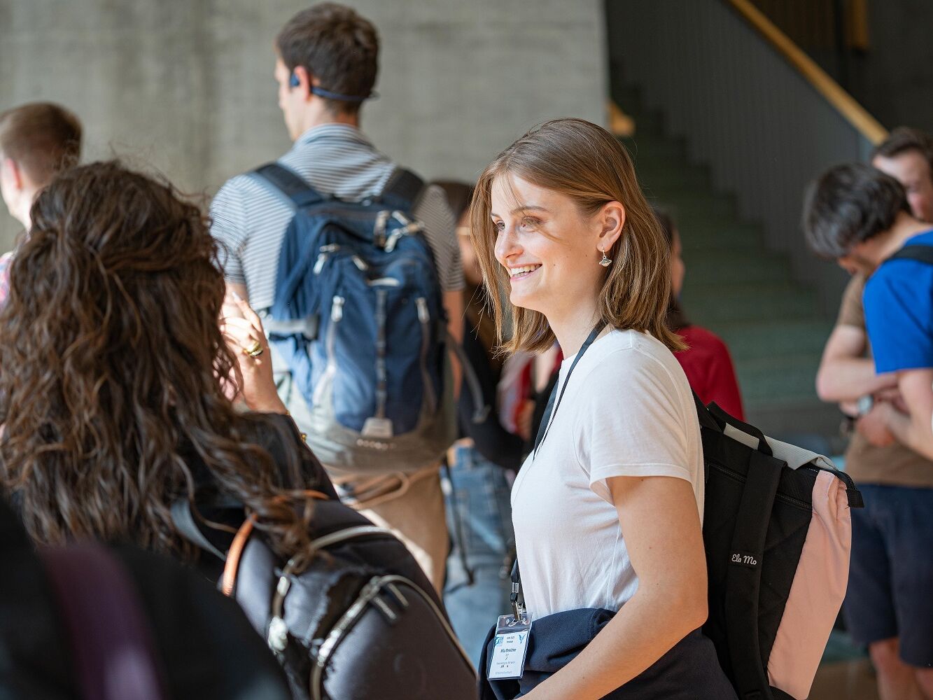 Junge Frau mit weißem T-Shirt und Namensschild lacht in einer Gruppe von Menschen, trägt einen Rucksack und steht in einem hellen Innenraum.