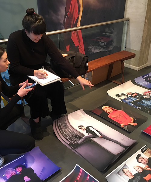 Zwei Frauen sitzen auf dem Boden einer Galerie und betrachten großformatige Fotografien, die vor ihnen ausgelegt sind. Eine Frau hält ein Handy, die andere macht Notizen in ein Heft und zeigt auf ein Bild. Es wirkt wie die Vorbereitung oder Planung einer