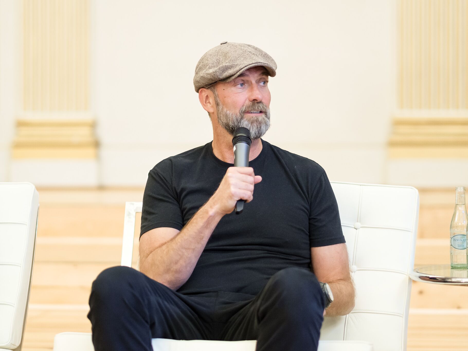 Ein Mann mit Bart, schwarzem T-Shirt und Schirmmütze sitzt auf einem weißen Stuhl, hält ein Mikrofon und schaut zur Seite.
