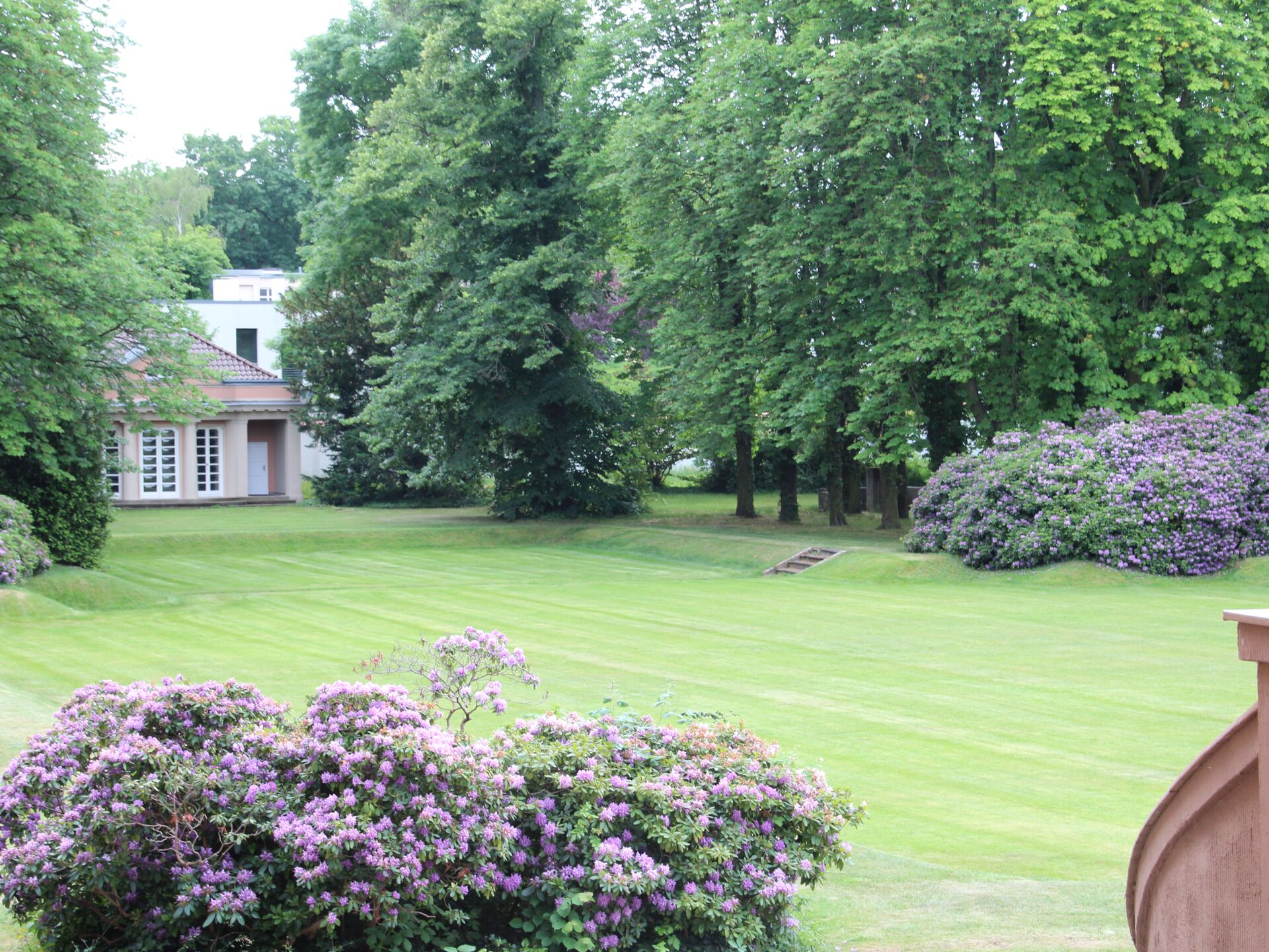 Gepflegter Garten mit blühenden Sträuchern, großer Rasenfläche und Bäumen neben einem Haus.