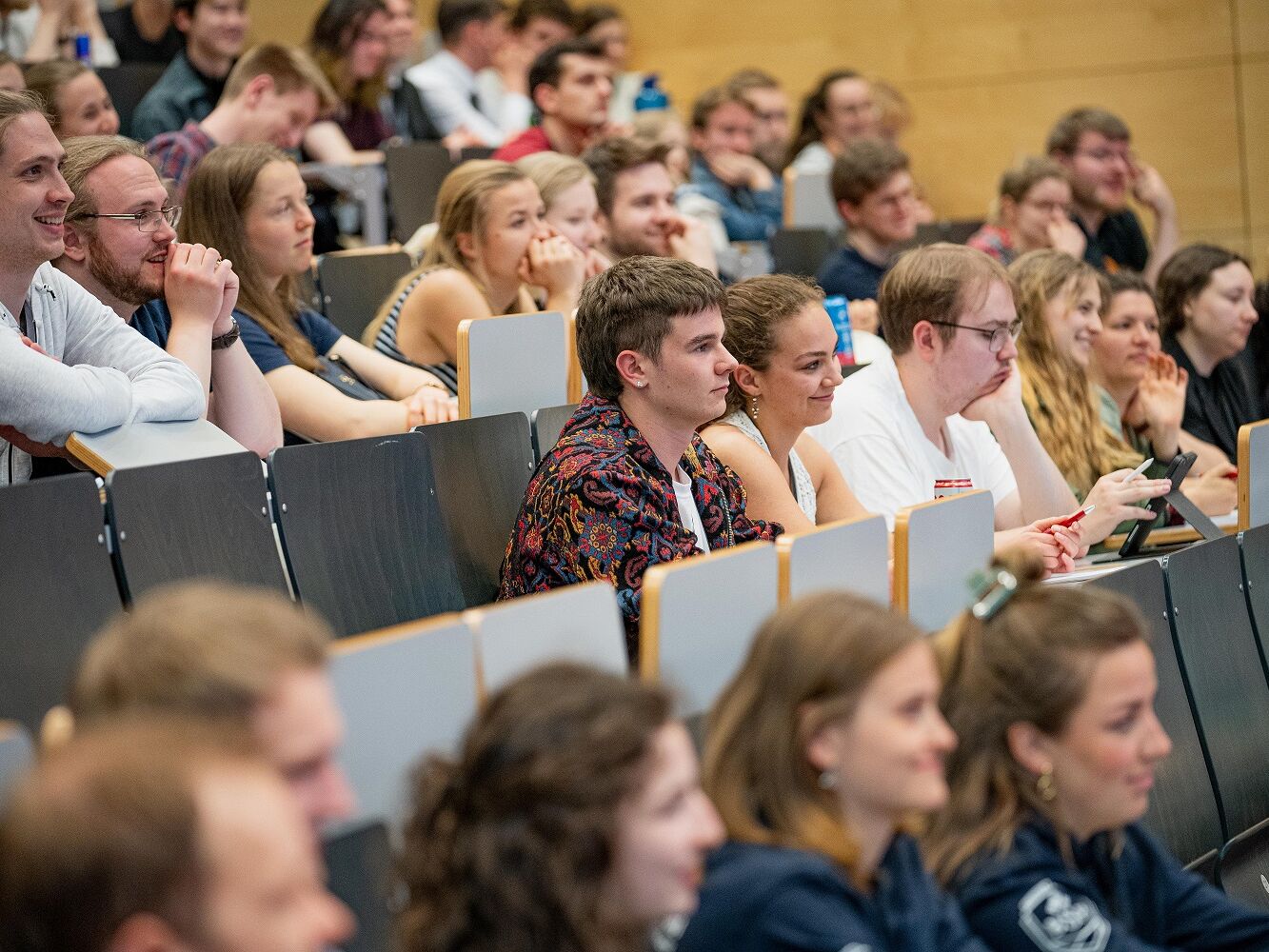 Viele junge Menschen sitzen dicht nebeneinander in einem Hörsaal. Sie hören aufmerksam zu, einige lächeln, andere machen sich Notizen.