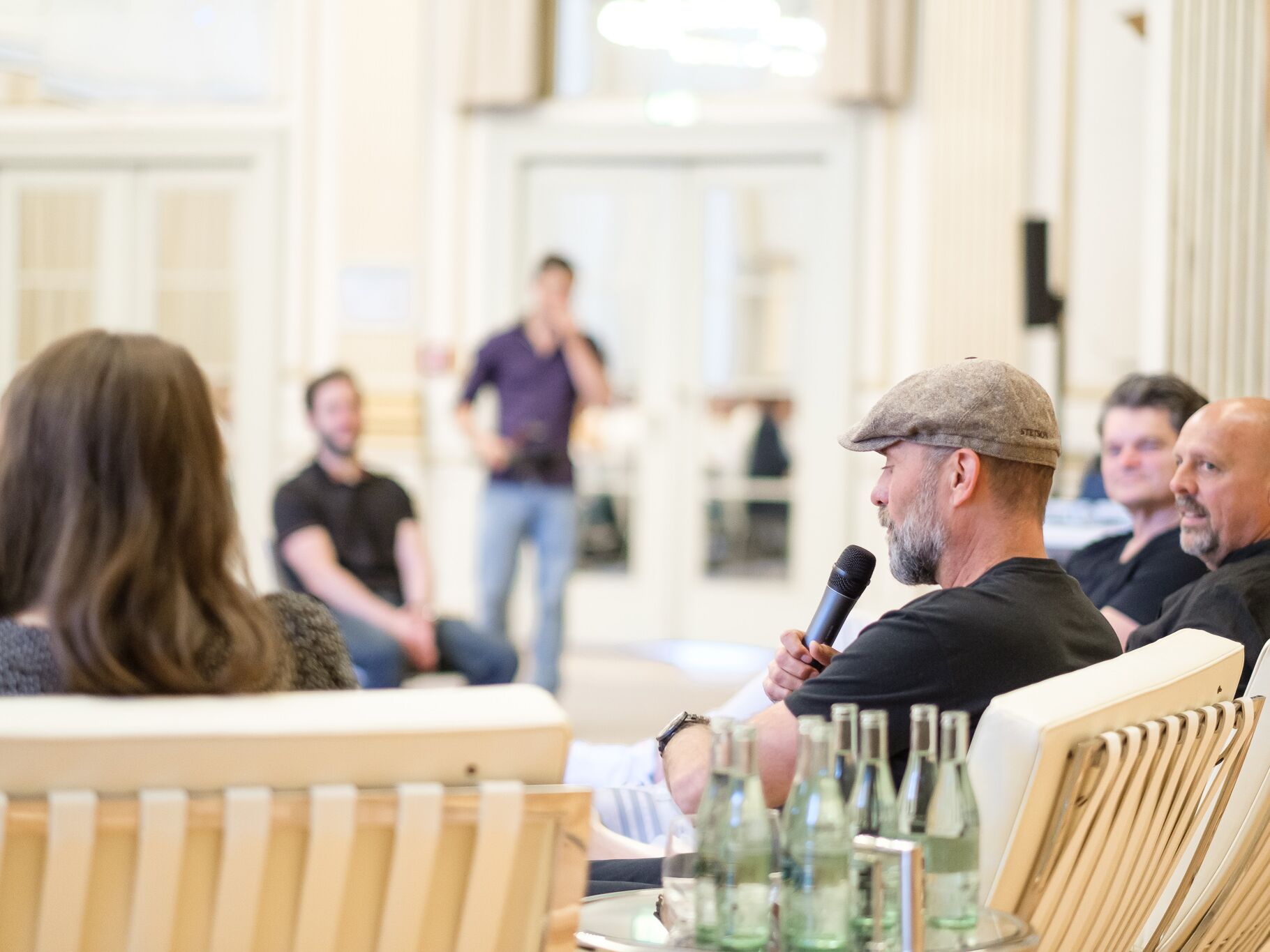 Mehrere Personen sitzen in weißen Sesseln in einem Saal. Ein Mann mit Schirmmütze und Bart spricht ins Mikrofon, andere hören zu. Im Vordergrund sind Wasserflaschen auf einem Tisch zu sehen.