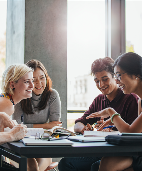 Gruppe junger Menschen sitzt zusammen an einem Tisch, lacht und arbeitet an Büchern und Notizen.