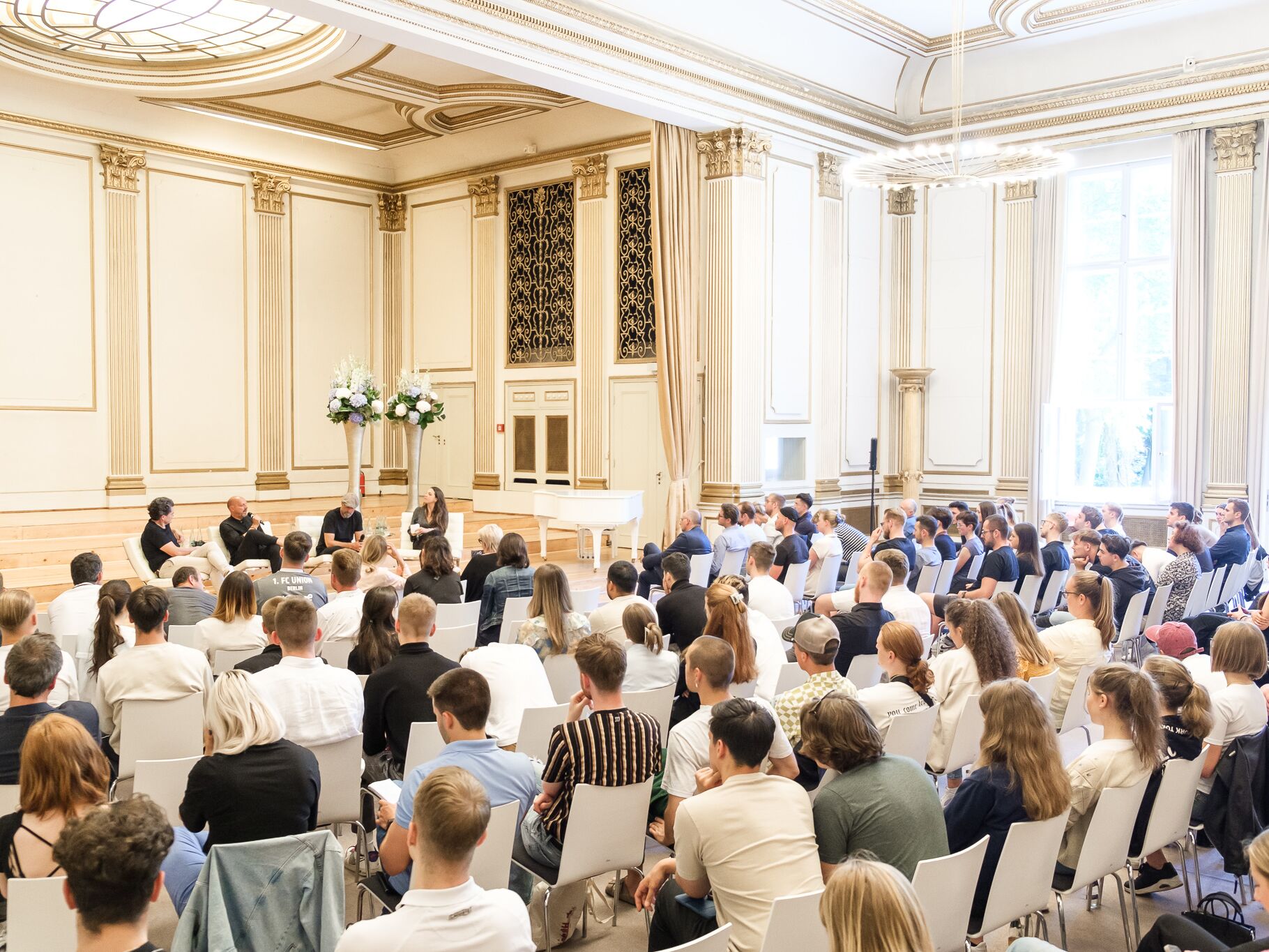 Zahlreiche Personen sitzen in einem prunkvollen Saal und verfolgen eine Podiumsdiskussion mit vier Rednern auf einer Bühne.