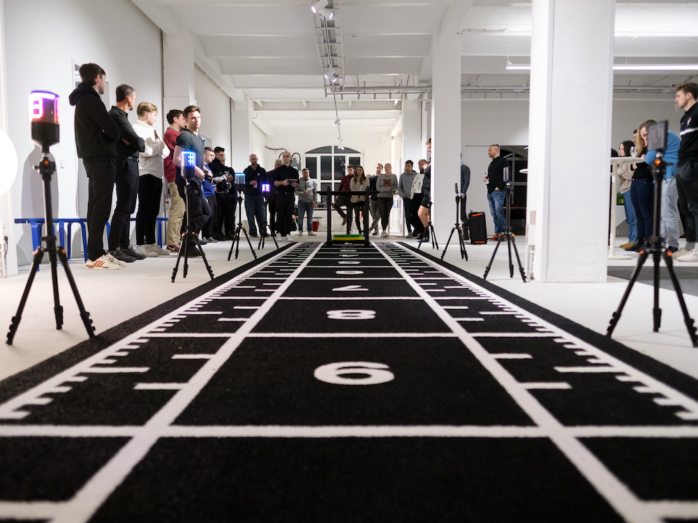Menschen stehen in einer Halle neben einer schwarzen Laufbahn mit Startmarkierungen und Messgeräten.