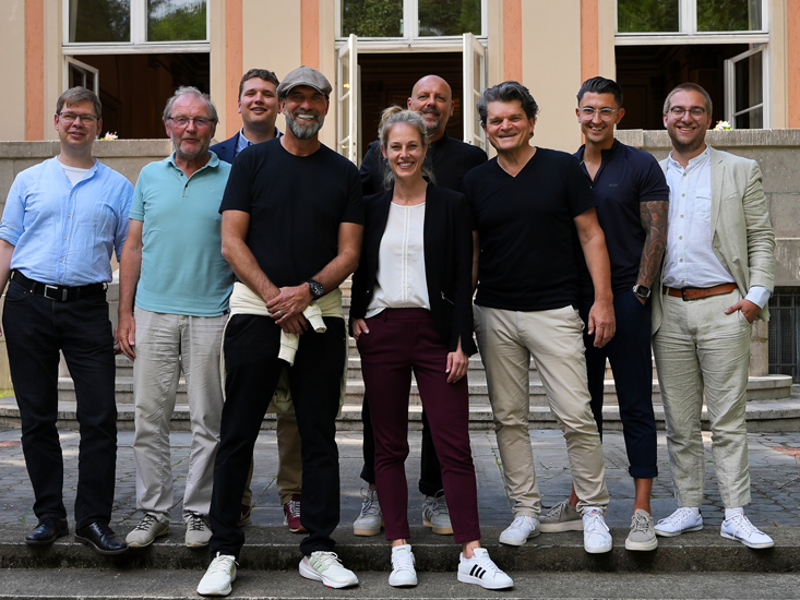 Eine Gruppe von neun Personen posiert gemeinsam draußen vor einem Gebäude und lächelt in die Kamera.