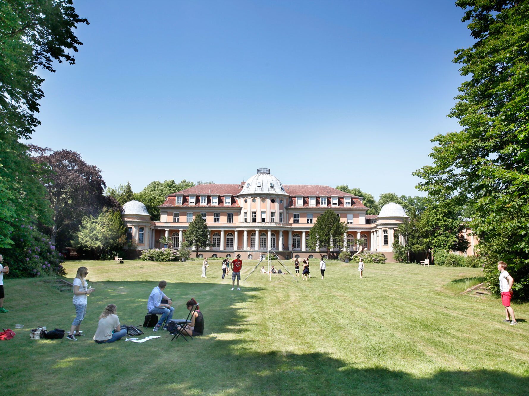 Großes historisches Gebäude mit Kuppeldach und Garten, in dem Menschen auf der Wiese sitzen und spielen.