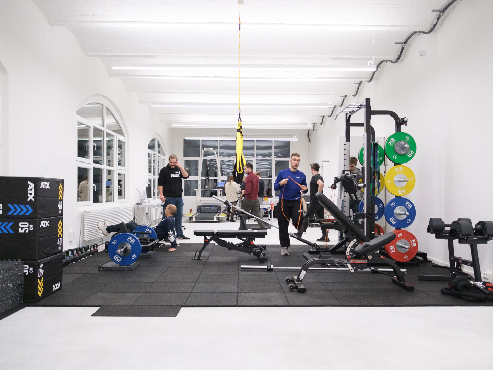 Menschen trainieren und unterhalten sich in einem modernen Fitnessraum mit Gewichten und Trainingsgeräten.