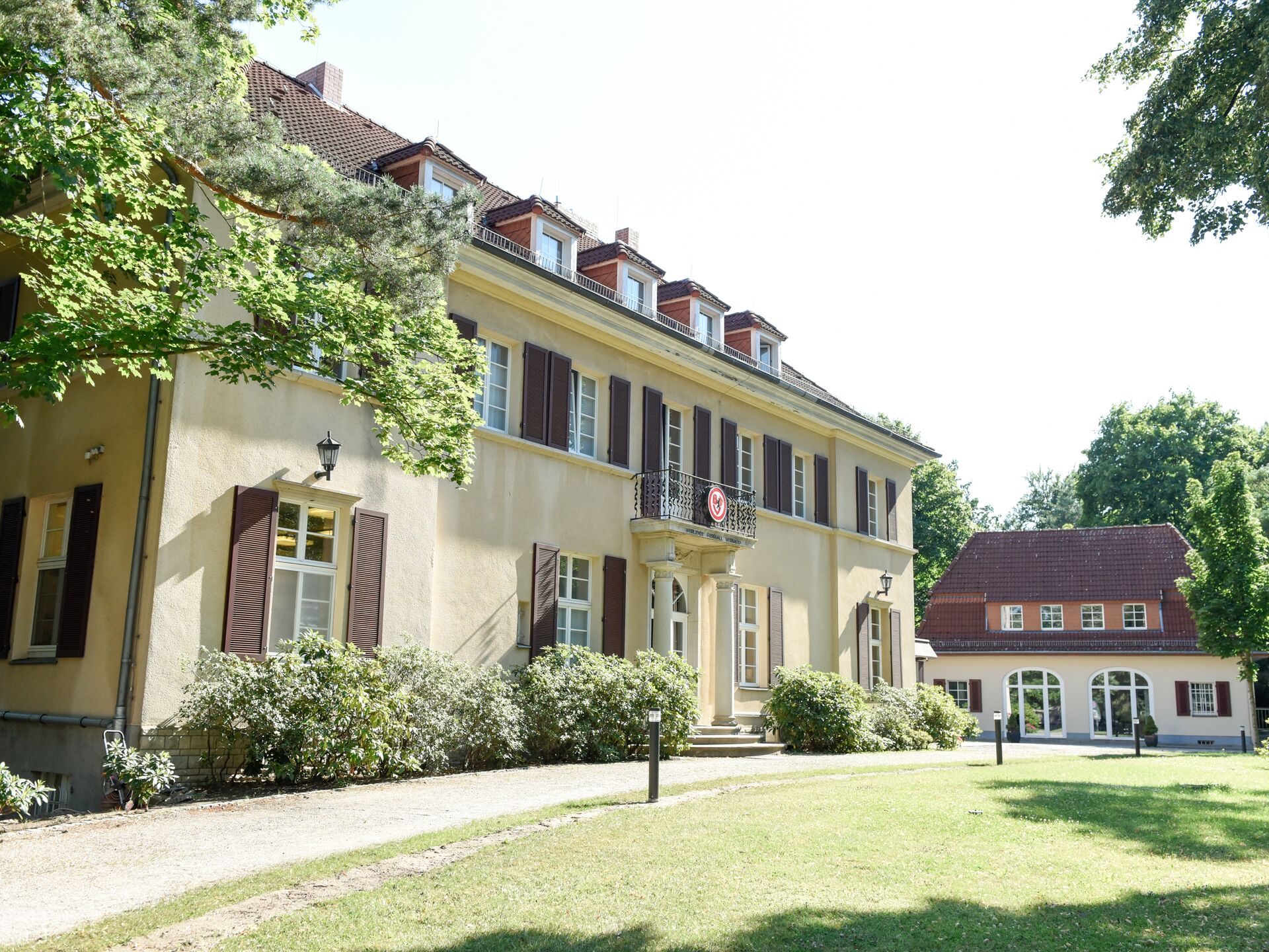 Großes helles Gebäude mit braunen Fensterläden und Balkon, umgeben von Garten und Nebengebäude.